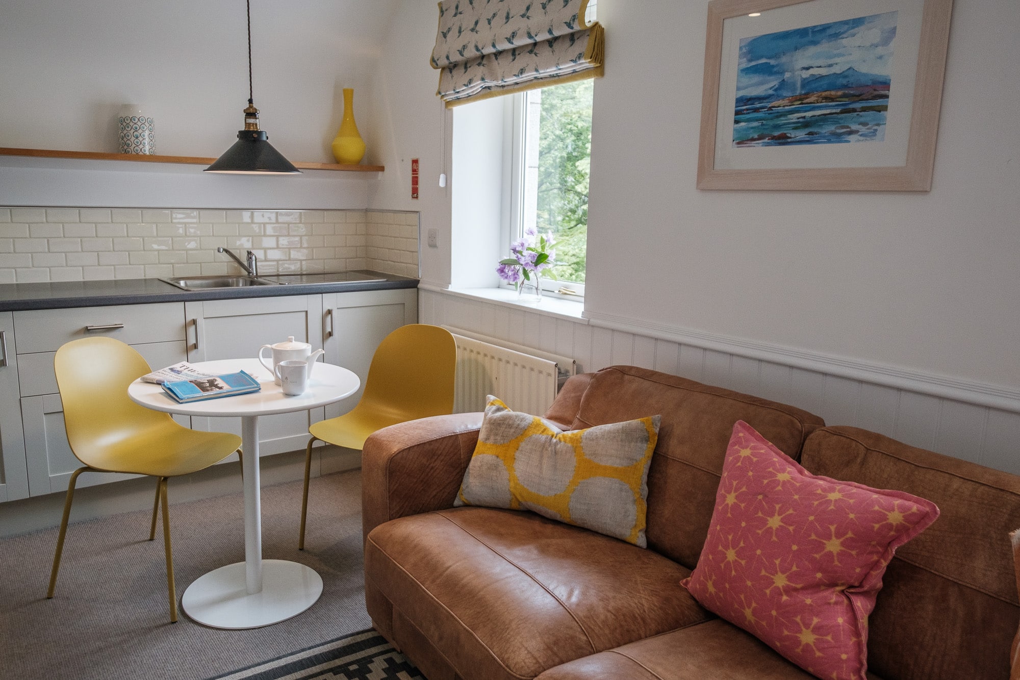 Open-plan kitchen and living area in Casa 24 with tan leather sofa, yellow chairs and coastal artwork at Douneside House